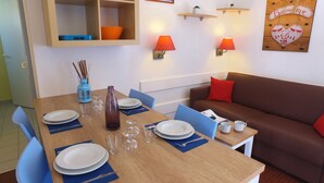 Ein Essbereich mit einem Holztisch, gedeckt für vier Personen, blauen Stühlen und einem braunen Sofa mit roten Kissen.
