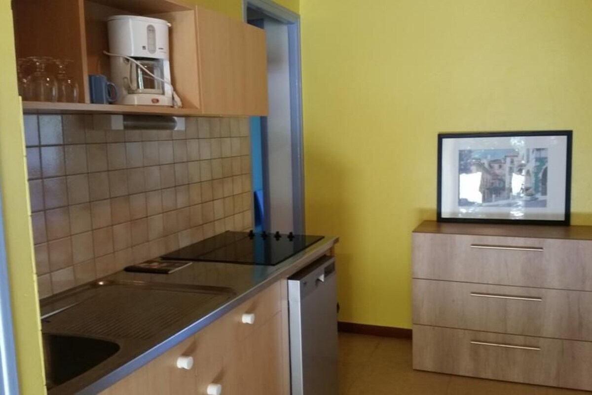A compact kitchen with a stove, sink, and a coffee maker on the countertop. There is a framed picture on a cabinet and a small cabinet with drawers.