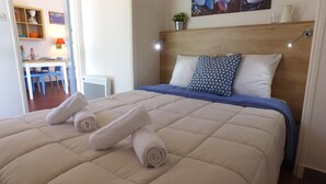 Ein ordentlich bezogenes Bett mit einem blau-weißen Kissen, einer beigen Steude und zwei aufgerollten Handtüchern.