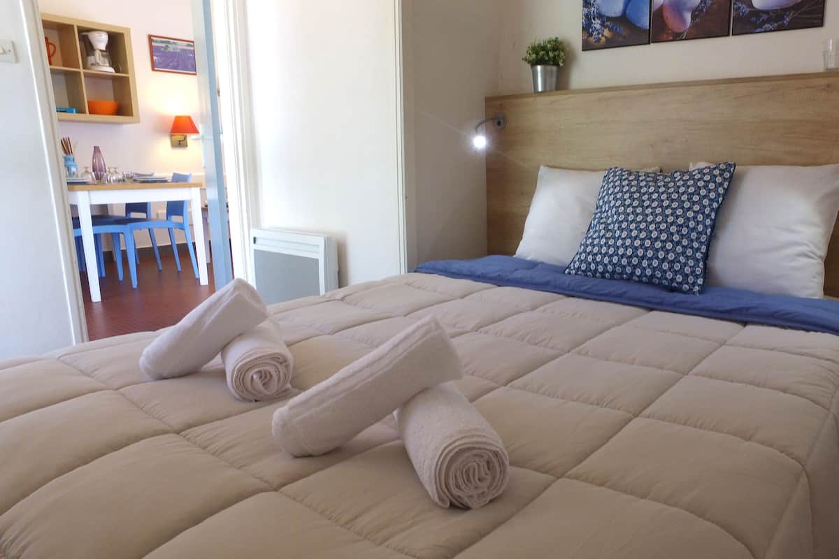 A neatly made bed with a blue and white patterned pillow, a beige quilt, and two rolled towels on the bed.