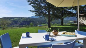 Ein Tisch mit Schachbrett, einer blauen Tasse und einer Schale mit Kiefernzapfen, draußen mit schöner Aussicht auf Hügel und einem Haus im Hintergrund.
