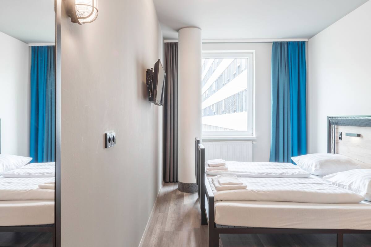 Habitación de hotel con dos camas, televisión, ventana con cortinas y suelo de madera.