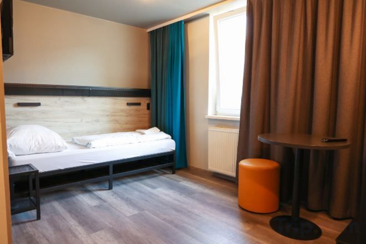 Ein Einzelbett mit Holzgestell, ein kleiner Tisch, ein orangefarbener Hocker und ein Fenster mit Vorhängen.