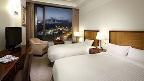 A hotel room with two beds, a desk with a television, a chair, and a large window with a city view. - The K-Hotel Seoul (Seoul)