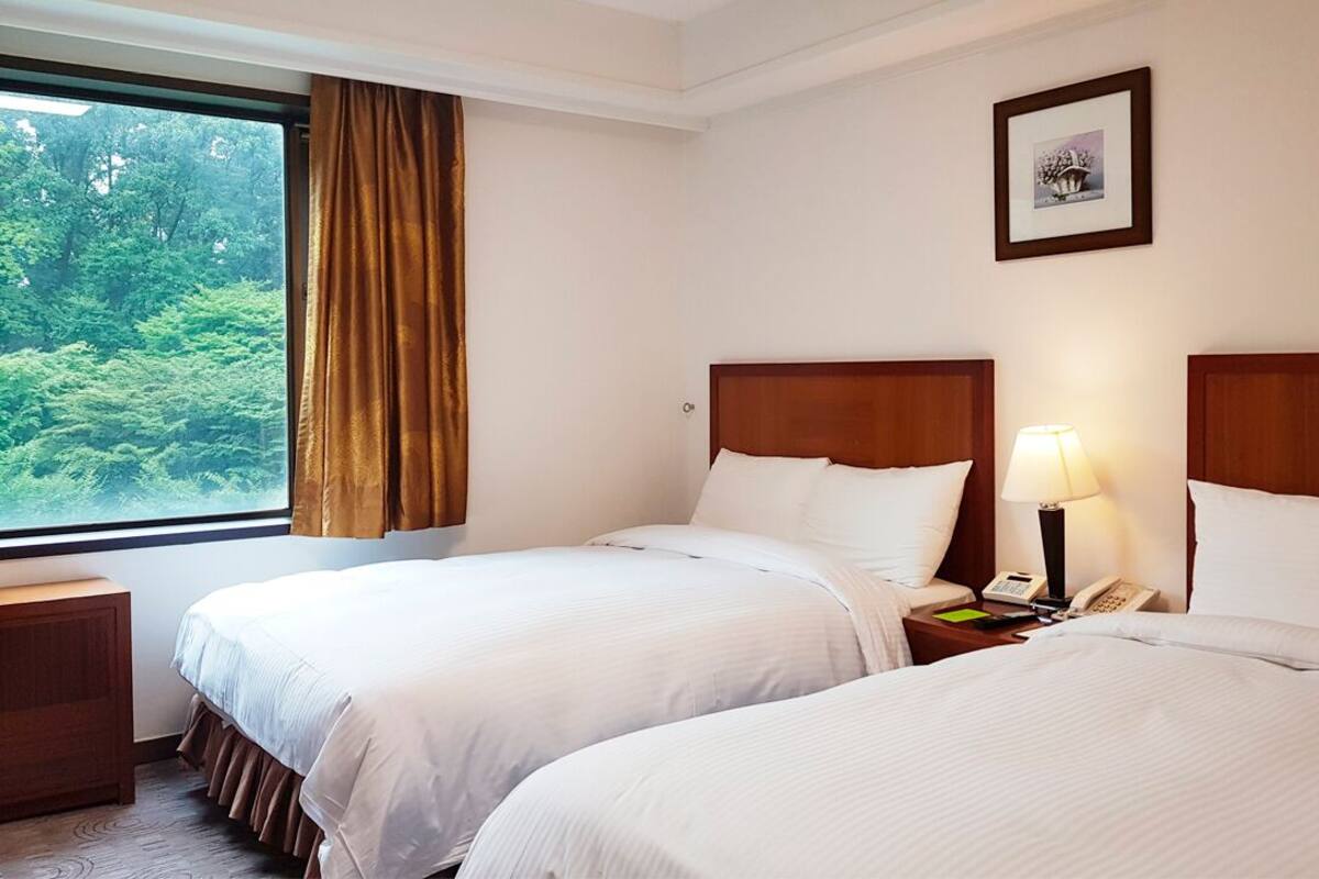 Una habitación de hotel con dos camas, una mesita de noche con lámpara, un teléfono y una ventana con cortinas.