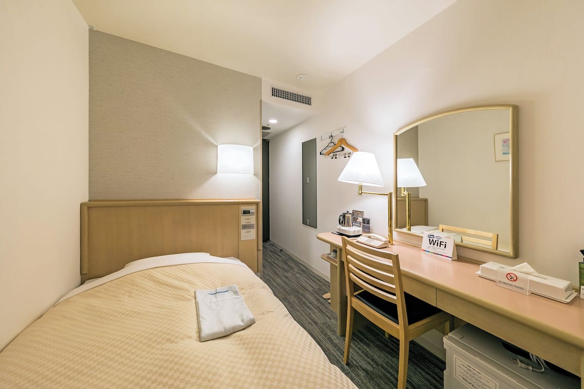 A hotel room with a bed, a desk, a chair, a lamp, a mirror, and a sign indicating WiFi availability.