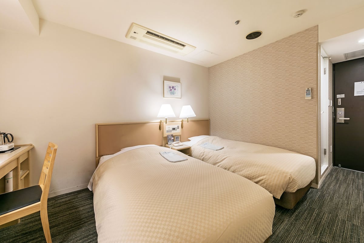 A hotel room with two beds, a desk, a chair, a nightstand with lamps, and an air conditioning unit.