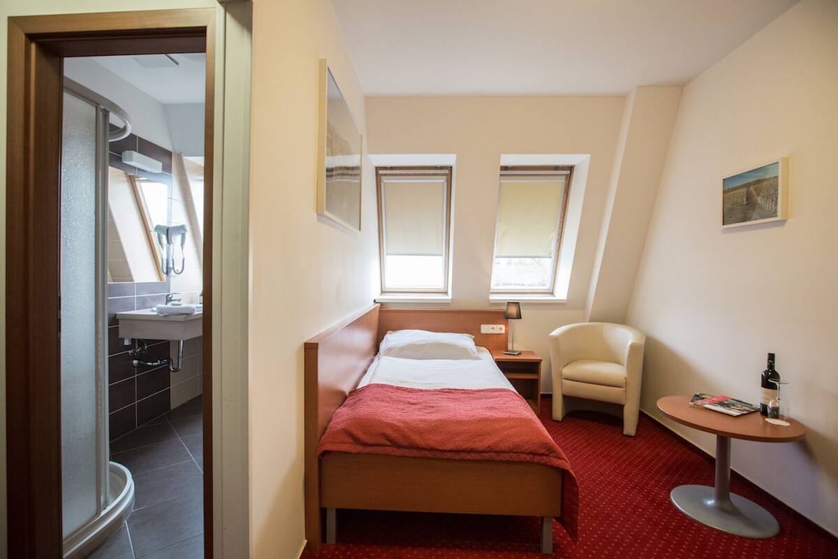 A compact hotel room with a single bed, a small desk, a chair, and a round table with a bottle and glass.