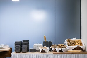 A buffet setup with stacked plates, bowls, a toaster, and baskets of bread and pastries. - Wine Wellness Hotel Amande (Hustopece)