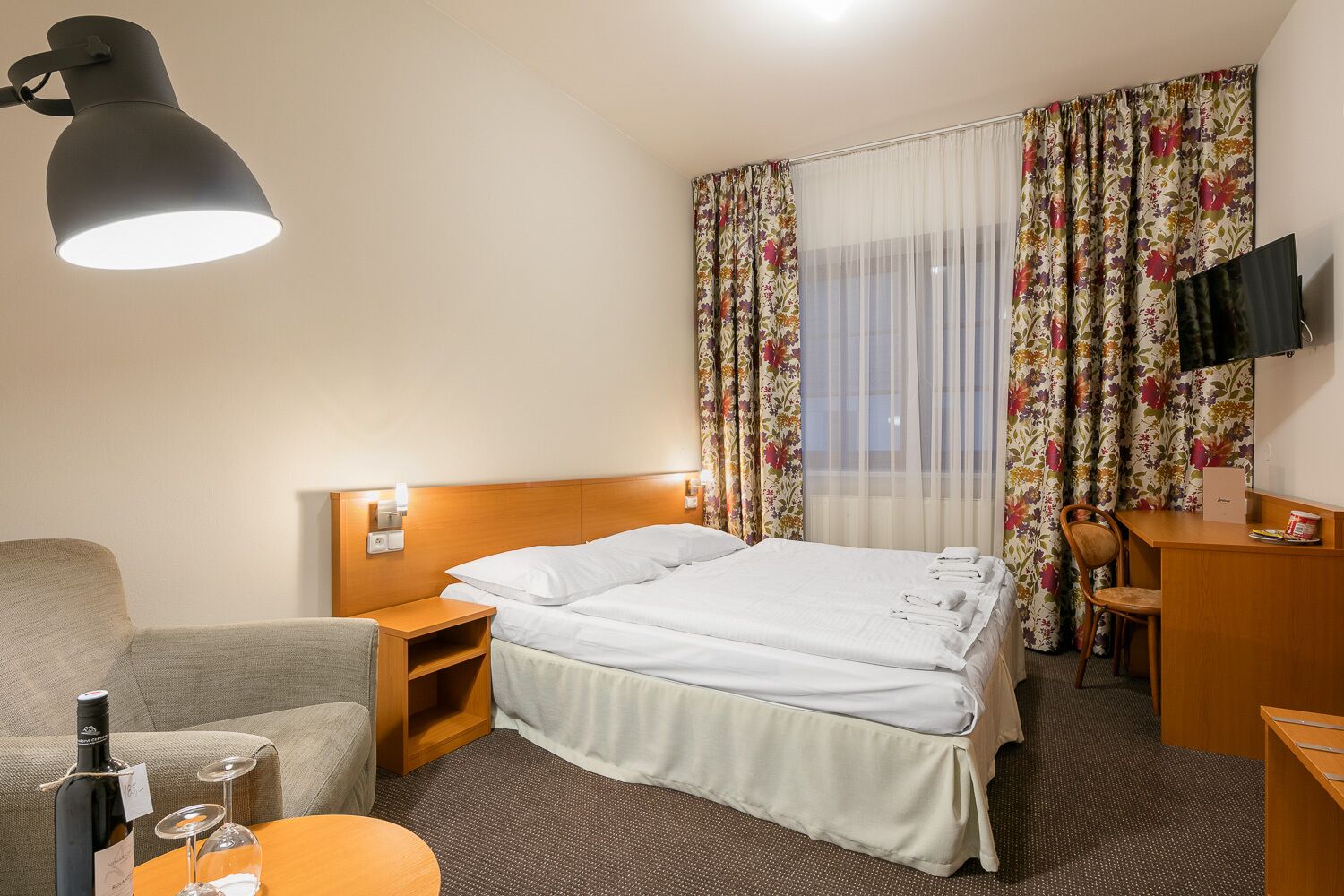 Habitación de hotel con cama, escritorio, silla, mesita de noche, lámpara, televisión y ventana con cortinas florales.