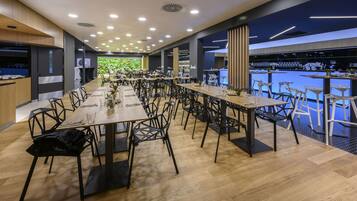 A modern dining area with long tables and chairs, a kitchen area on the left, and a bar area on the right.