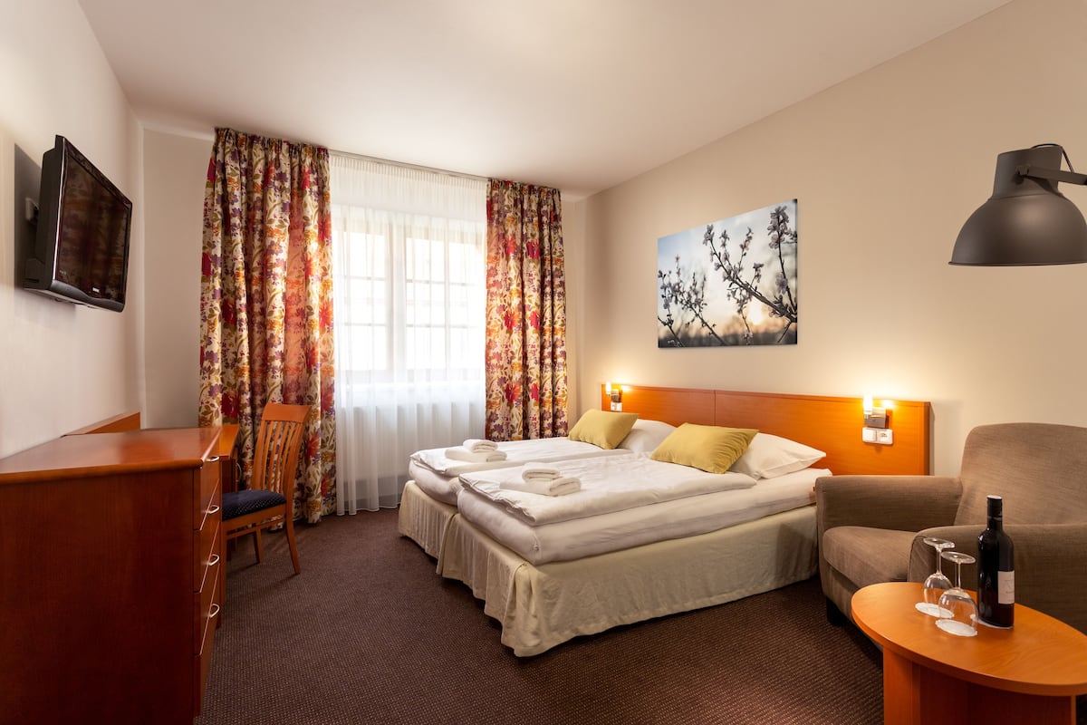 A hotel room with a bed, a desk, a chair, a sofa, a table with a bottle and glasses, and a painting on the wall.