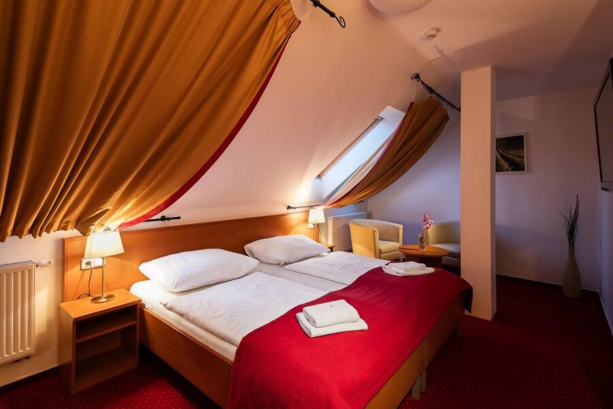 A hotel room with a bed, two bedside tables, a lamp, a chair, a small table, a radiator, and a window with curtains.
