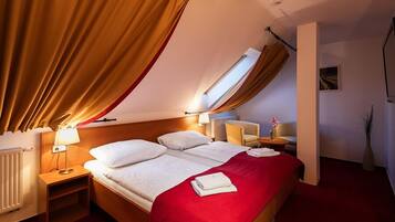 Habitación de hotel con cama, dos mesitas de noche, lámpara, silla, mesa pequeña, radiador y ventana con cortinas.