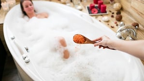 Una persona en una bañera llena de burbujas, con una mano sosteniendo una cuchara de madera con una sustancia roja.