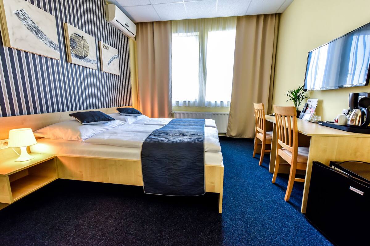 A hotel room with a bed, bedside table with lamp, a desk with chairs, a TV, and a window with curtains.