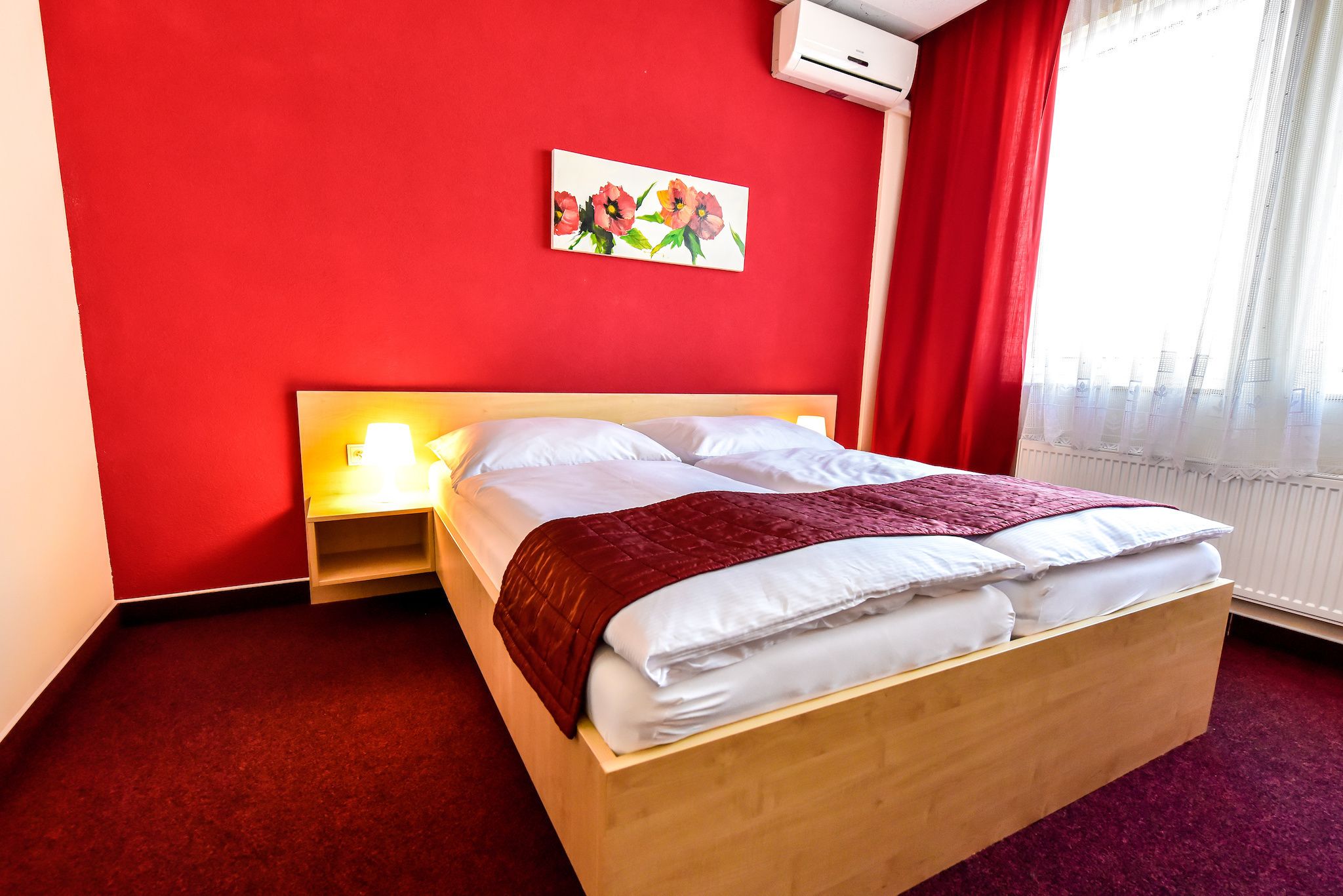 A neatly made bed with white linens and a red blanket, a bedside table with a lamp, a red wall with a floral painting, and a window with sheer curtains.