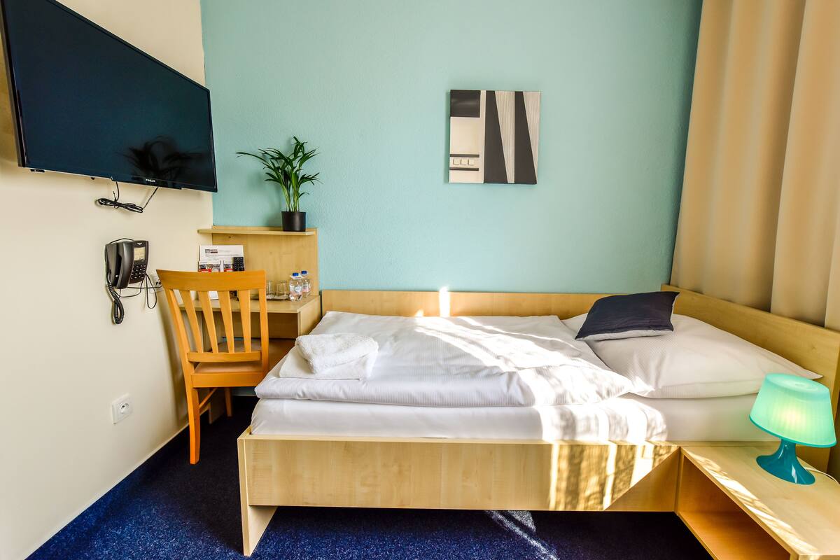 A hotel room with a bed, a desk with a chair, a telephone, a plant, and a painting on the wall.