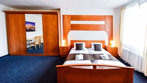 A hotel room with a wooden bed, two bedside tables with lamps, a large window with curtains, and a door leading to another room. - City Hotel (Brno)