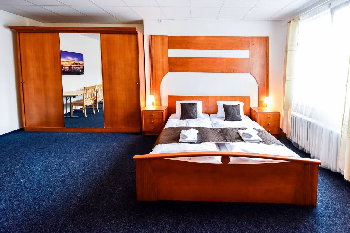 A hotel room with a wooden bed, two bedside tables with lamps, a large window with curtains, and a door leading to another room.