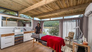 A room with a wooden ceiling, a bed with a red blanket, a wooden desk with a chair, a large window with curtains, and a view of a lake.