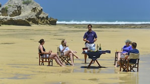 A group of people seated on beach chairs on a sandy beach with a rocky outcrop in the background and the ocean in the distance.
