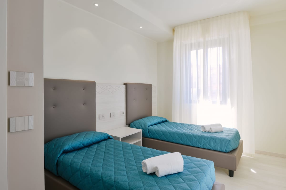 A hotel room with two beds, each with a teal quilted bedspread and a white towel on one bed.