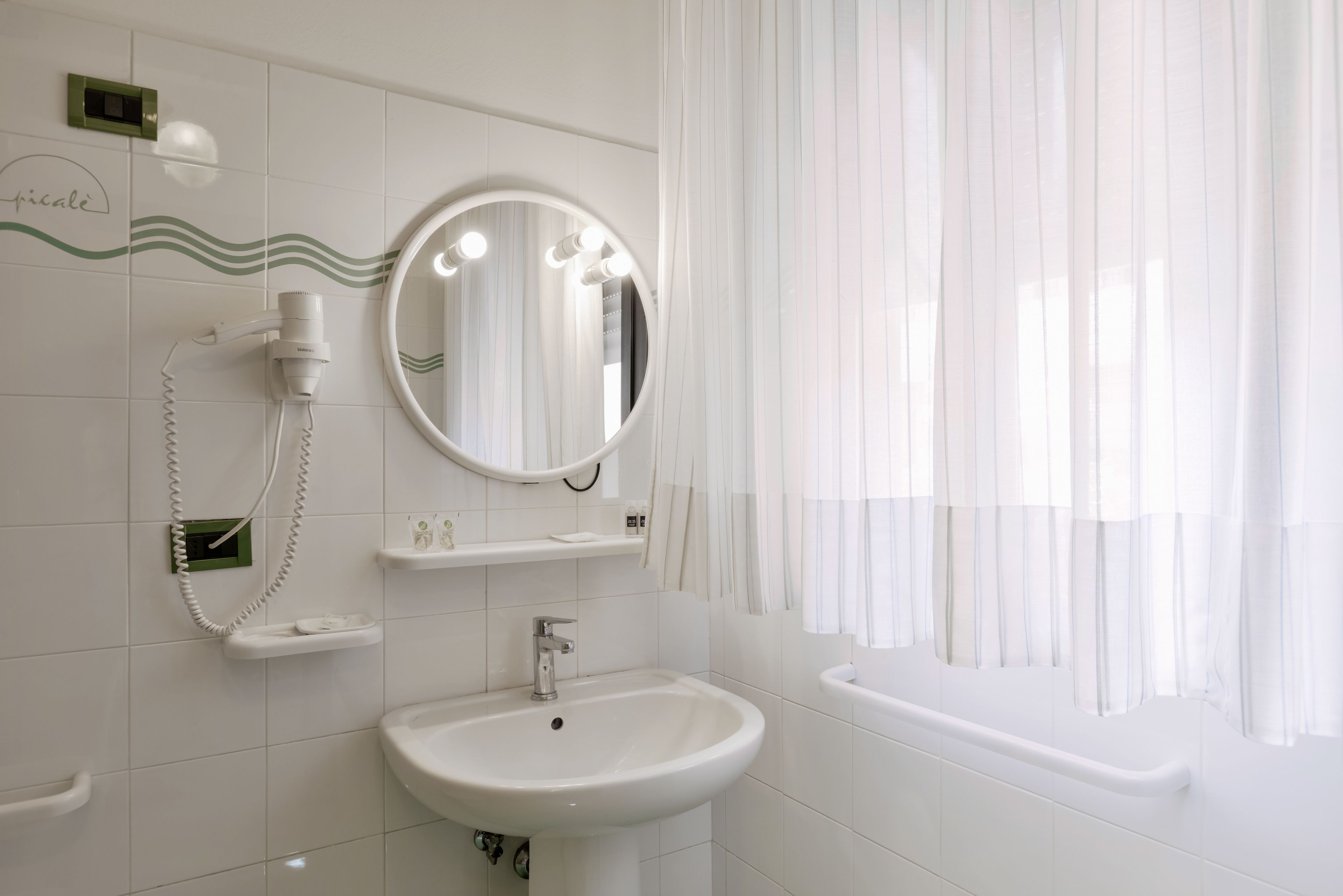 Un bagno con un lavandino bianco, uno specchio e un asciugacapelli. Si vede una finestra con tende leggere.