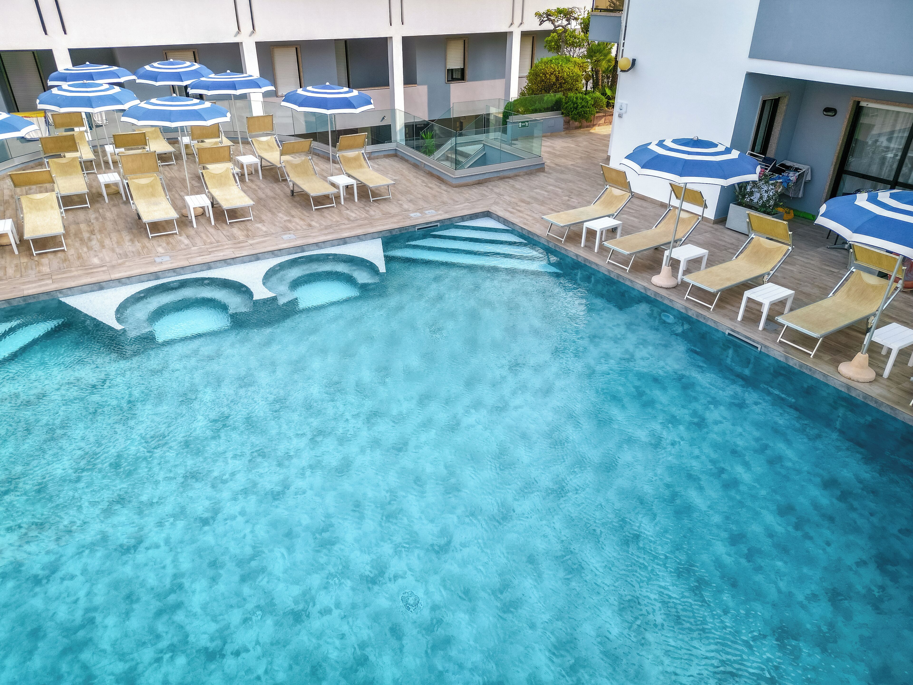 A pool area with multiple sun loungers and umbrellas, surrounded by a glass railing and a building facade.
