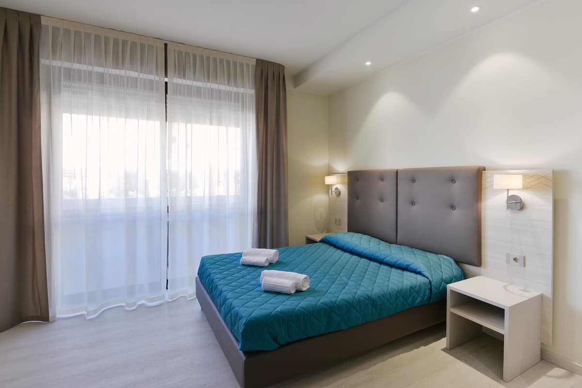 A modern hotel room with a large bed, a bedside table, and a window with sheer curtains.