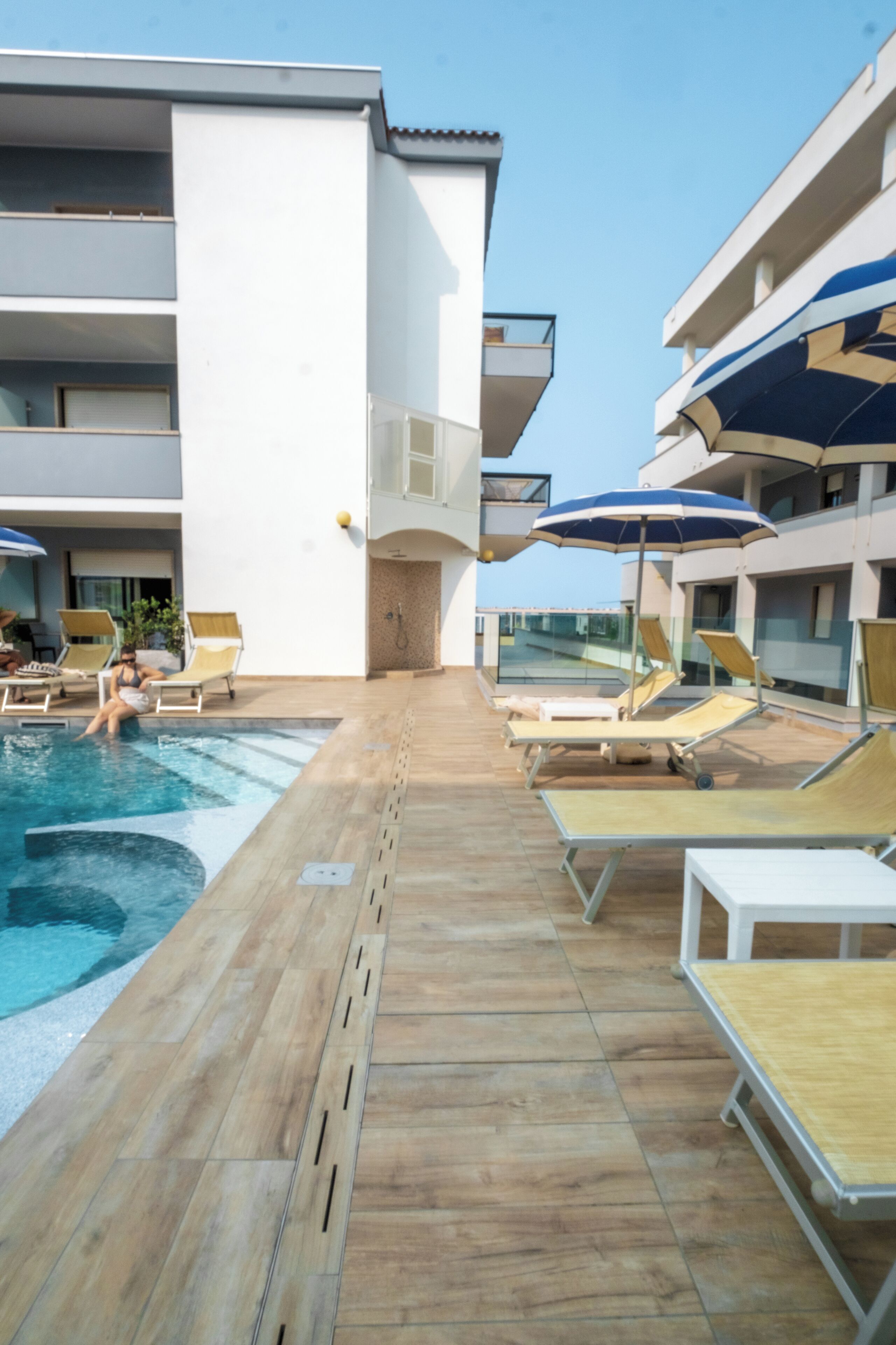 A pool area with lounge chairs and umbrellas, a clear sky, and a building with balconies.