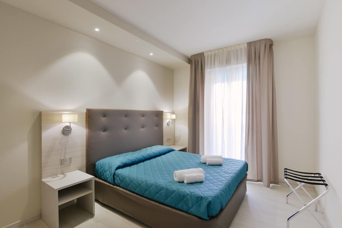 A modern hotel room with a large bed, bedside tables, a nightstand, a chair, and a window with curtains.