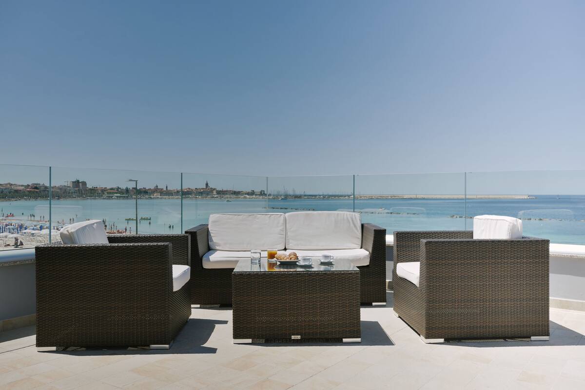A balcony with wicker furniture and a glass table, overlooking a beach and the sea.