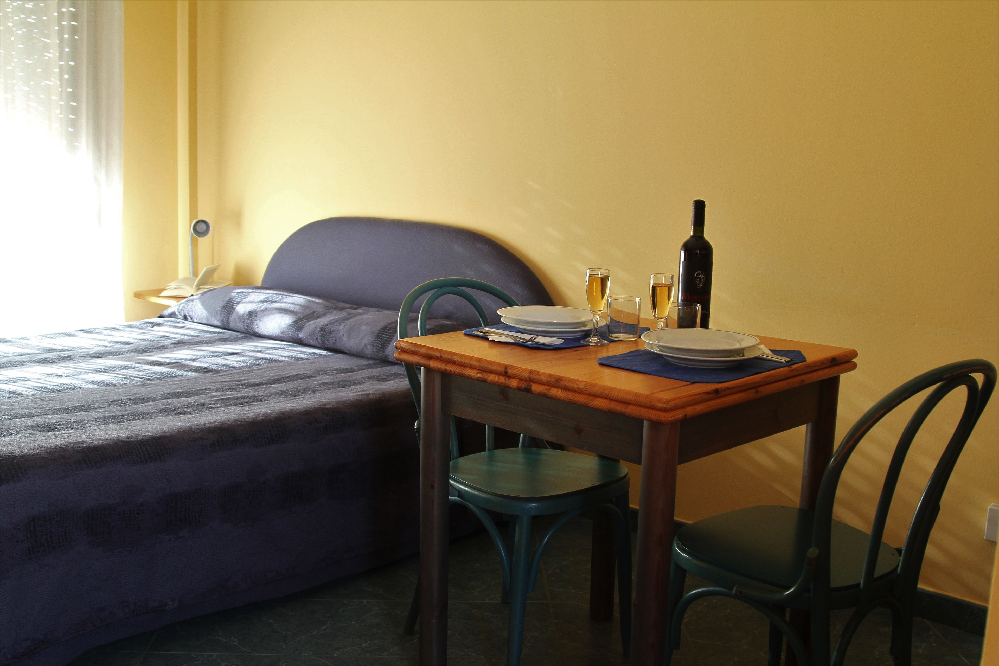 A room with a bed, a small wooden table with a bottle, glasses, and plates, and a chair.