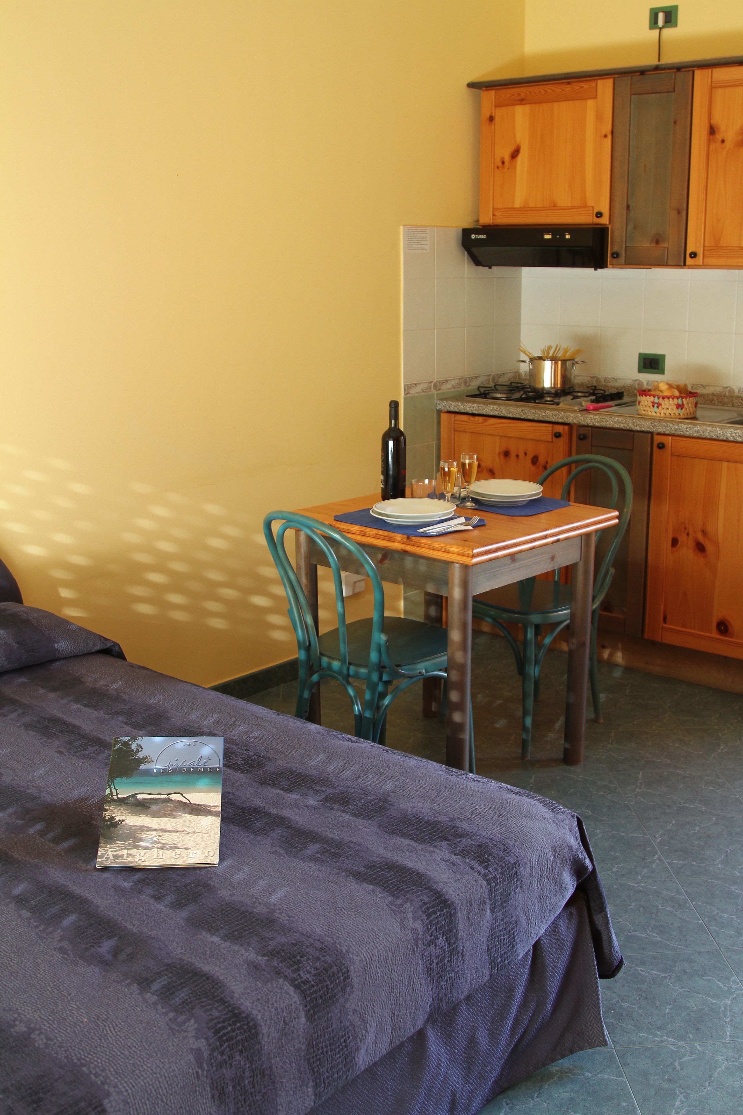 Ein Zimmer mit einem Bett, einem Esstisch mit Stühlen, einer Küchenzeile mit Holzschränken und einer gefliesten Spritzschutzleiste.