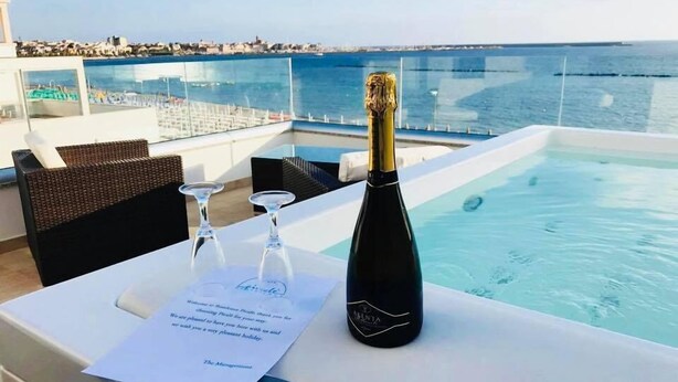 A table with a bottle of champagne, two glasses, and a menu on a terrace overlooking a pool and the sea. - Residence Picalè (Alghero)
