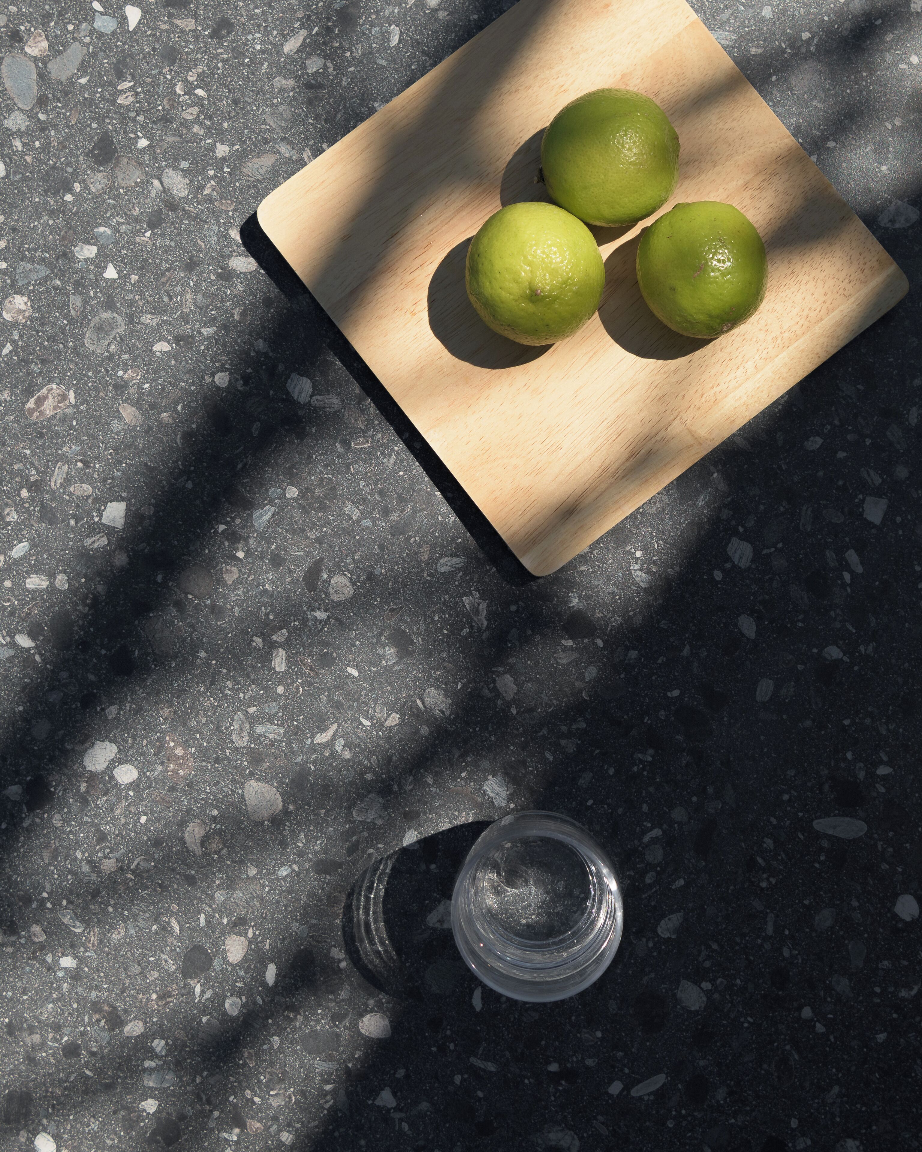 A wooden cutting board with three green limes on a speckled countertop.