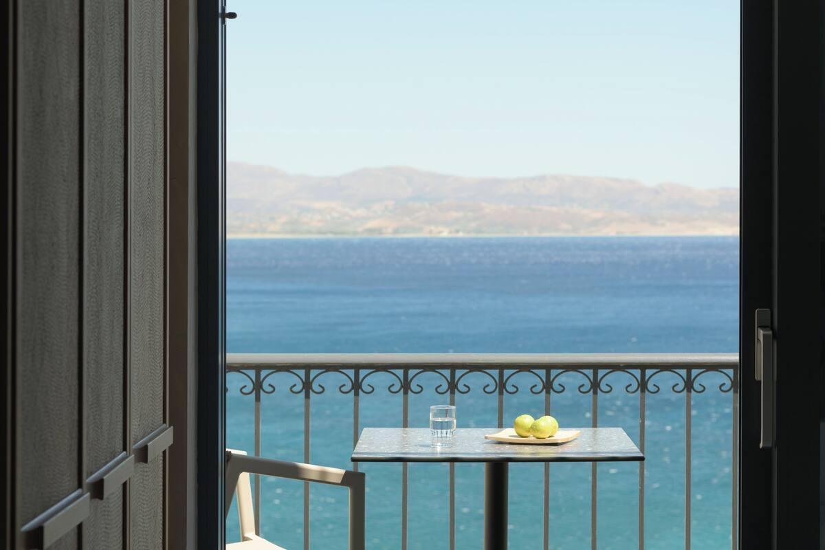A balcony with a table, chair, and a view of the sea and distant mountains.