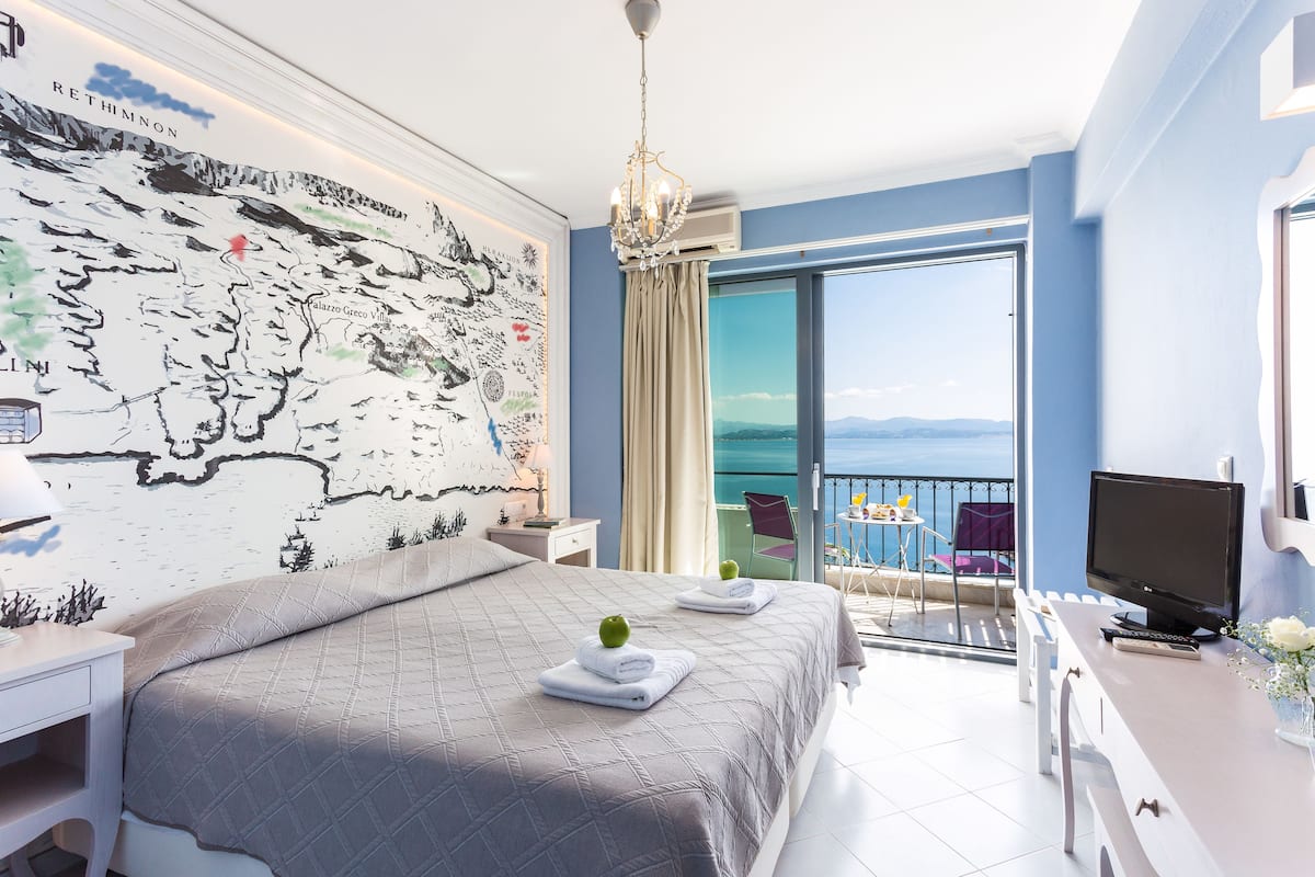 A hotel room with a large bed, a desk with a TV, a map on the wall, and a balcony with a view.