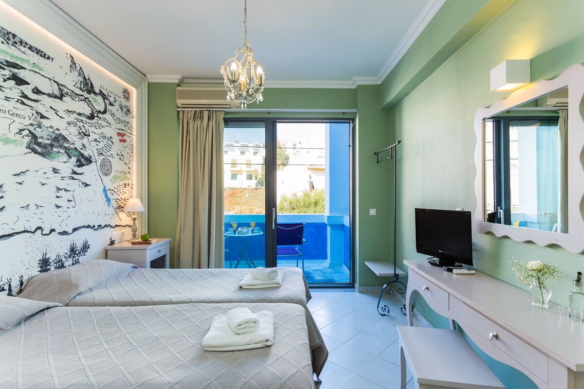 A hotel room with two beds, a television, a desk, and a balcony with a view of buildings.