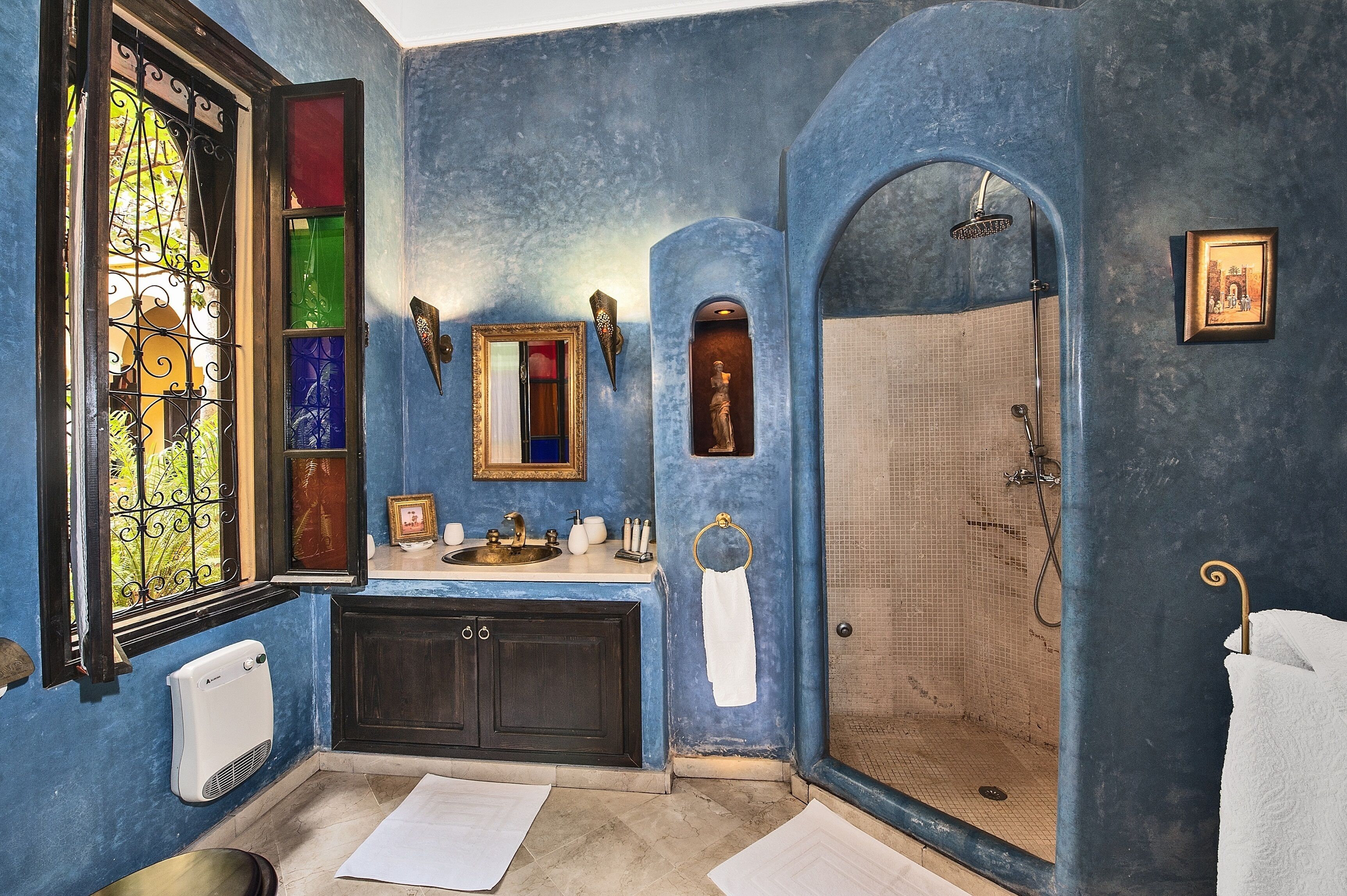 Un baño con pared azul, un ventanal grande con una reja de hierro decorativa, un lavamanos con un espejo encima y una zona de ducha.