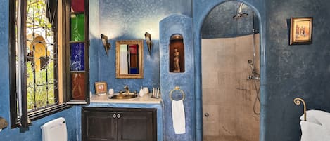 Un baño con pared azul, un ventanal grande con una reja de hierro decorativa, un lavamanos con un espejo encima y una zona de ducha.