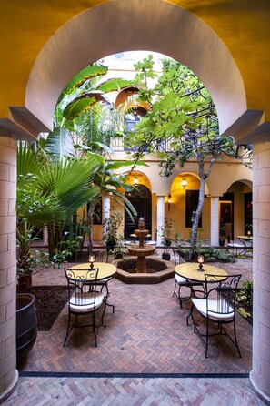 Un patio interior con una fuente, dos mesas redondas con sillas y plantas en macetas.