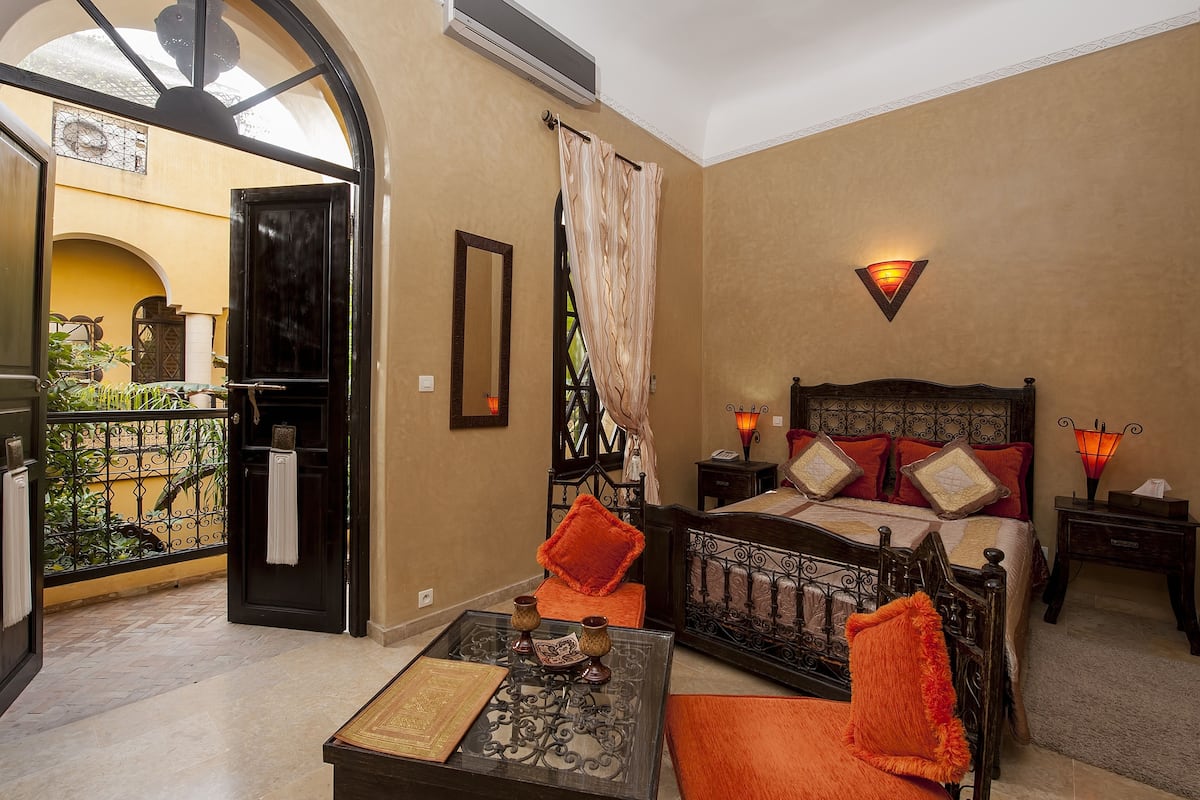 A room with a bed, a table with a book, a balcony with a view, and a wall-mounted light fixture.