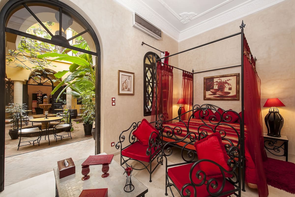 A room with a four-poster bed, red cushions, a table with a vase, and a view of an outdoor patio with a fountain and chairs.