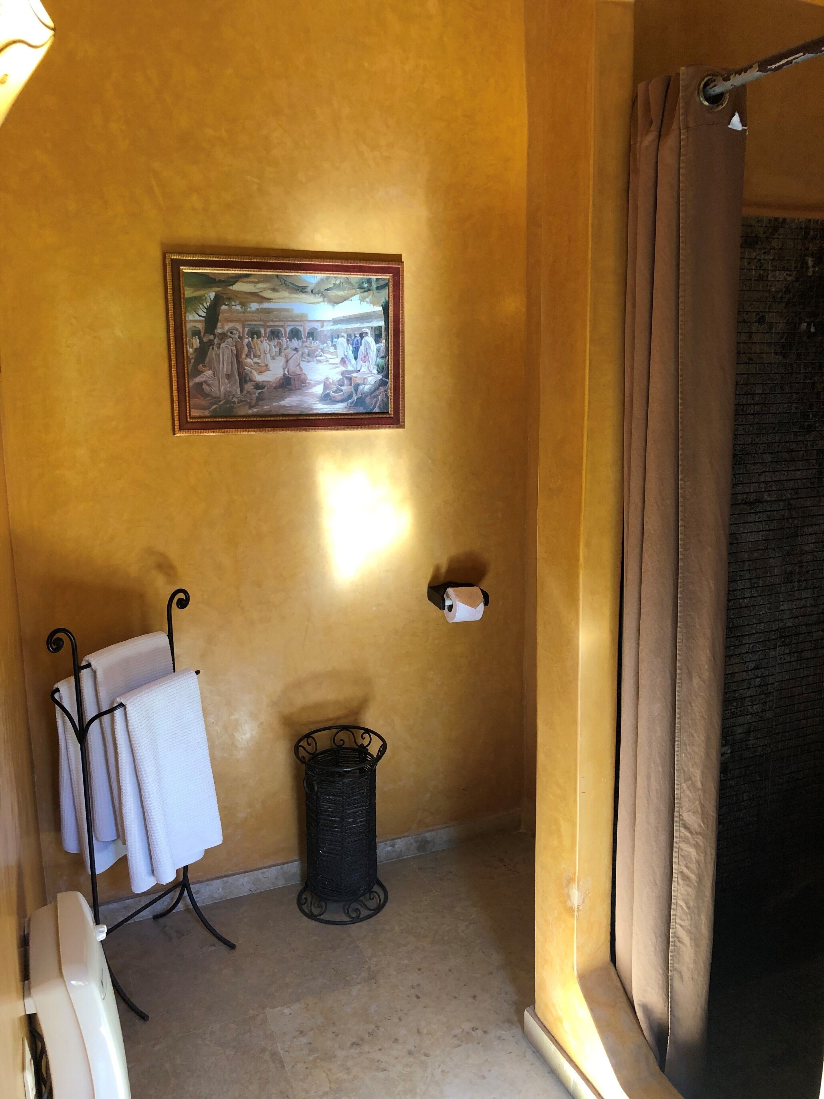 A bathroom with a yellow wall, a framed painting, a towel rack with towels, a trash bin, and a curtain.