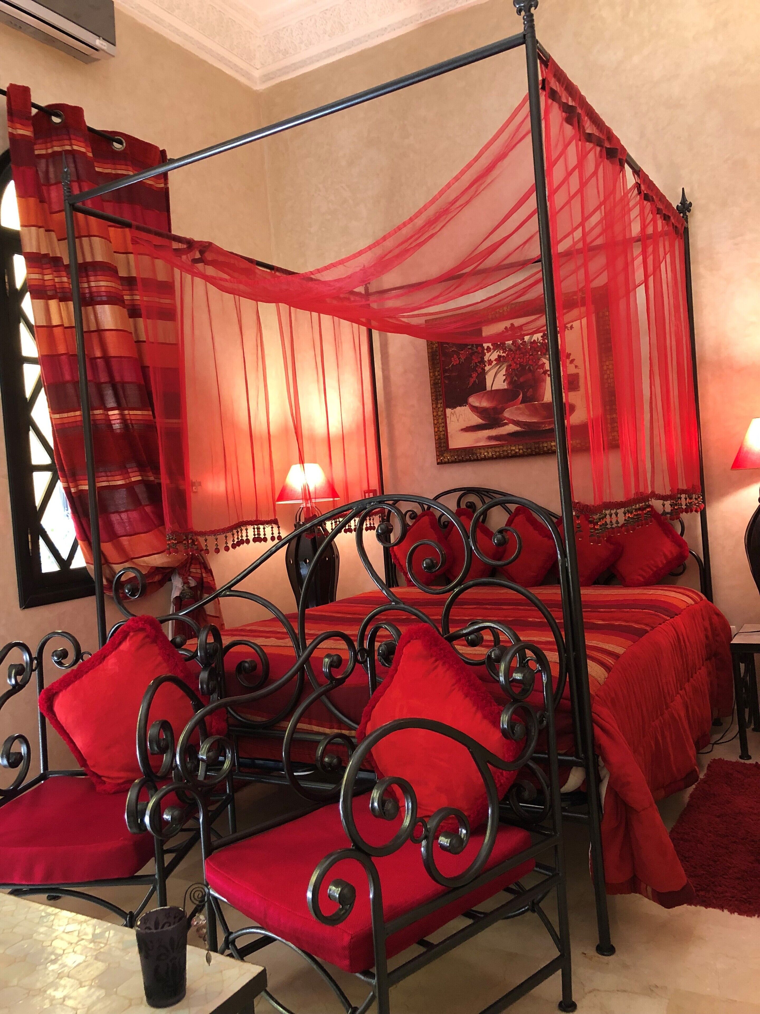 A four-poster canopy bed with red bedding and pillows, a red curtain, and a painting on the wall.