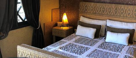 A hotel room with a bed, two chairs, a nightstand, an air conditioner, and a wall-mounted lamp.