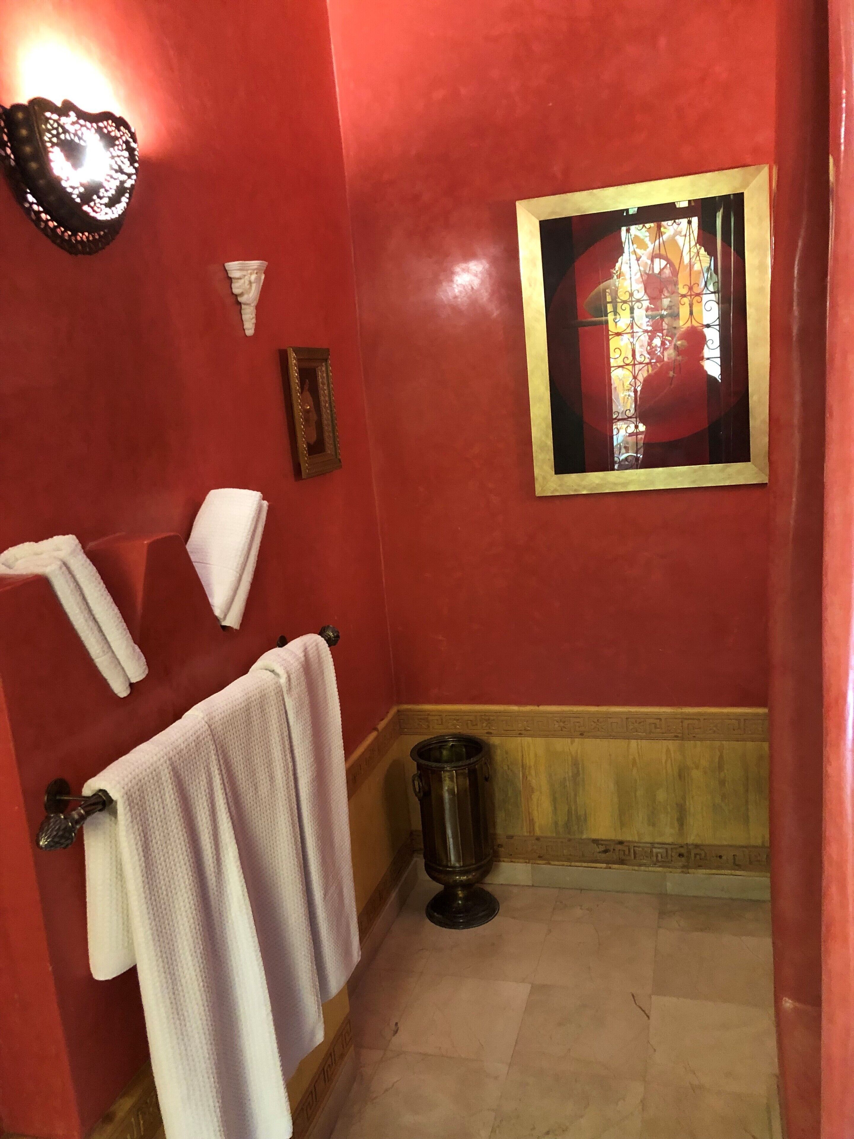 Un baño con paredes rojas, un perchero blanco, un cuadro enmarcado y un cubo de basura.