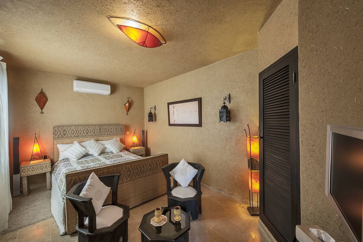A hotel room with a bed, two chairs, a small table with drinks, a TV, and decorative wall lights.
